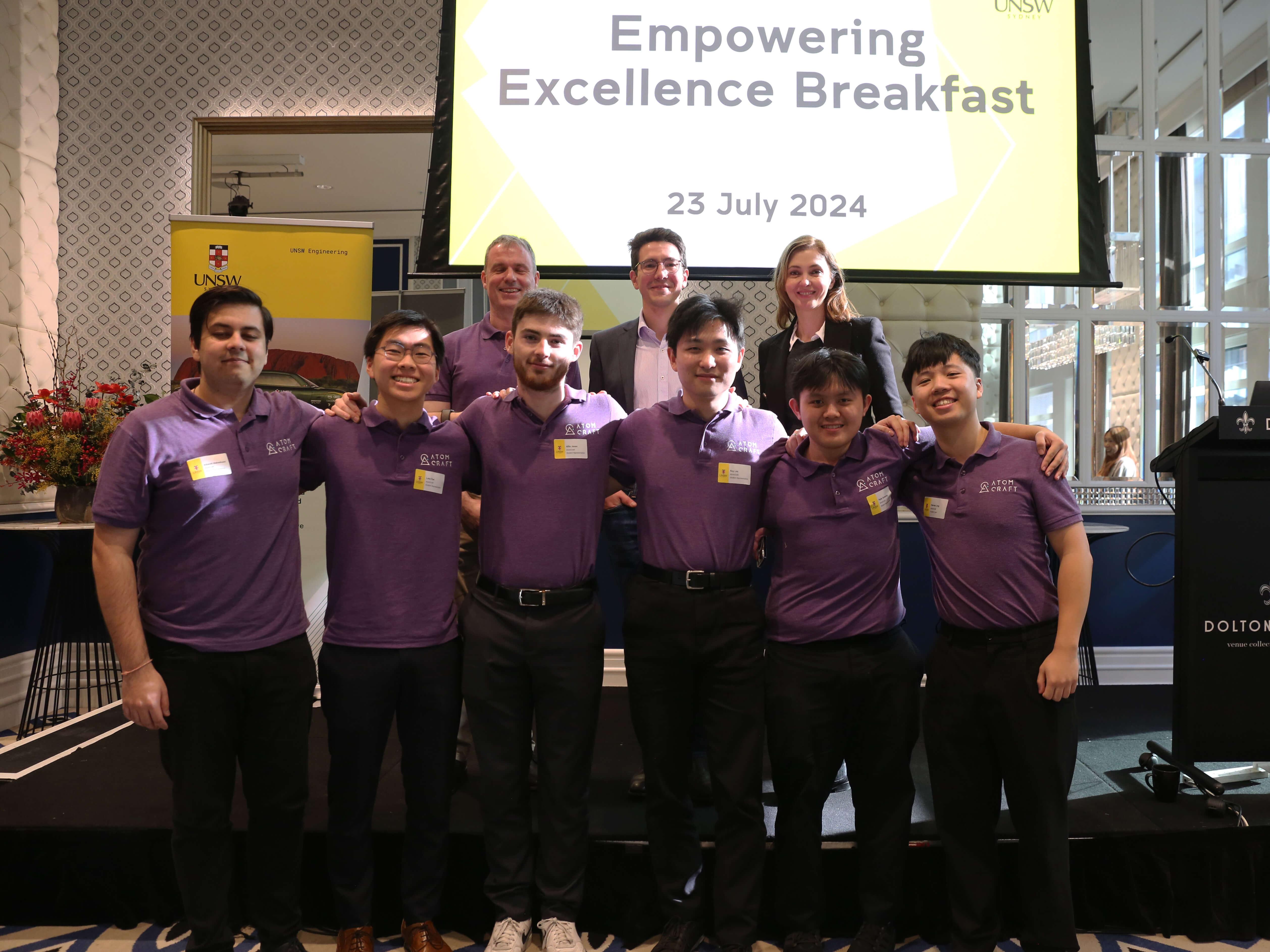 AtomCraft members with arms around each other's shoulders in front of a screen which reads 'Empowering Excellence Breakfast'
