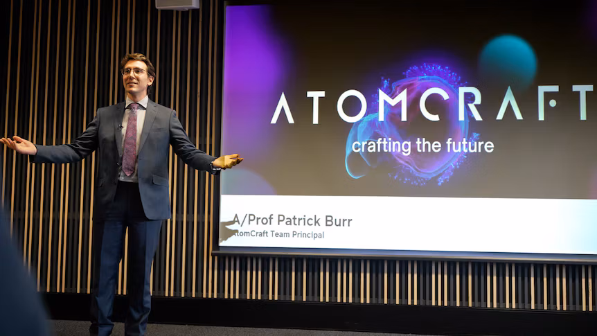 A man in a suit stands on stage, gesturing with his hands in front of a screen displaying the word 'Atomcraft'